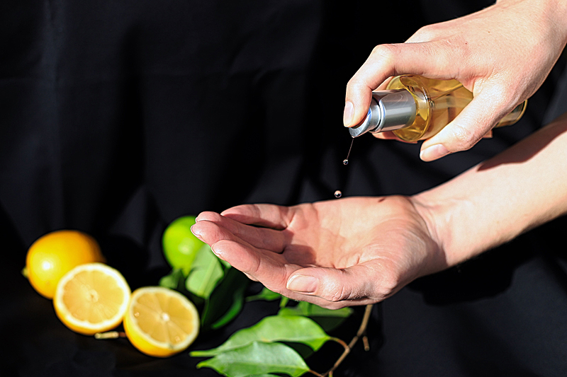 Das Bild zeigt eine Hand mit einer Körperöl-Dispenserflasche, die Körperöl in die Handfläche einer anderen Hand tropft. Im Hintergrund sind eine aufgeschnittene Zitrone, eine weitere Zitrone und eine Limette sowie grüne Blätter zu erkennen