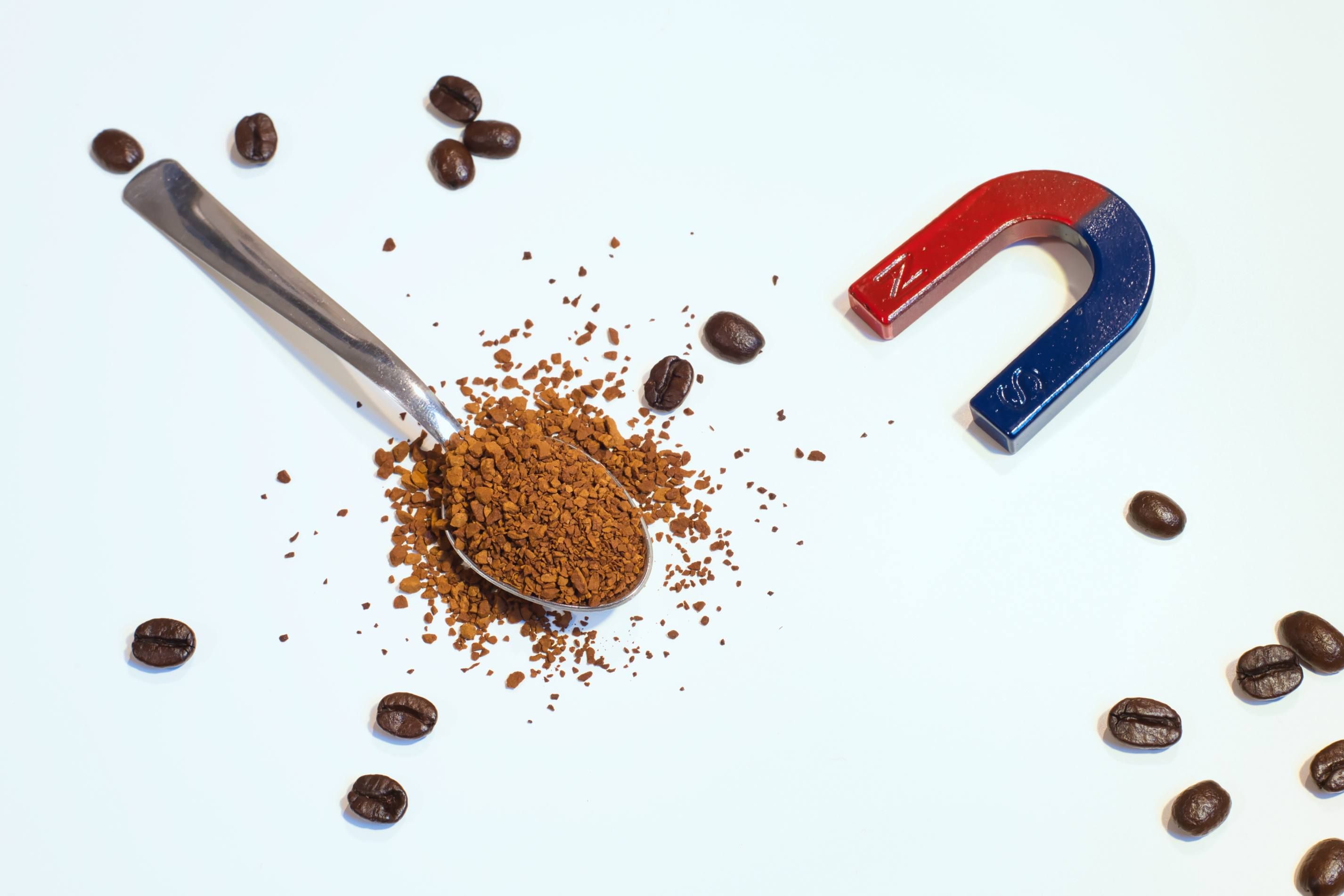 Zu sehen ist ein Teelöffel mit löslichem Kaffee in Granulatform, der von einigen ganzen Kaffeebohnen sowie einem hufeisenförmigen Magnet umgeben ist.