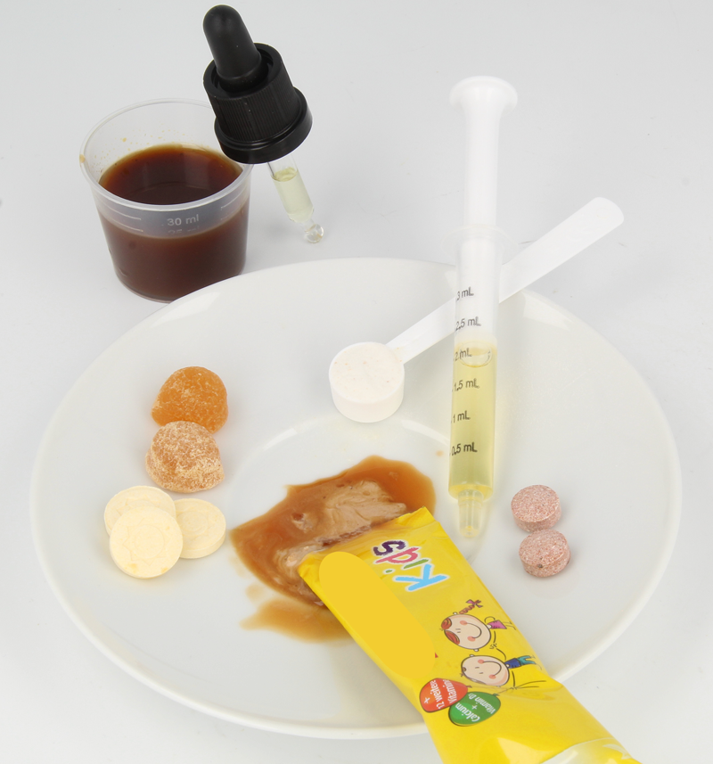 Zu sehen sind für Kinder vorgesehene Nahrungsergänzungsmittel in Form von Lutschtabletten, Fruchtgummis, Pulver in einem Dosierlöffel, einer Tropfpipette sowie Flüssigkeiten in einem Portionsstick, einem Dosierbecher und einer Dosierspritze.