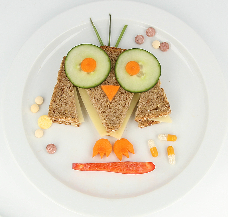 Zu sehen ist eine Brotzeit, die für Kinder optisch ansprechend in Eulenform angerichtet ist. Auf dem Teller liegen außerdem Nahrungsergänzungsmittel in verschiedenen Darreichungsformen.