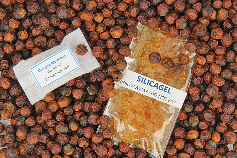 Dargestellt sind ein weißes Sauerstoffabsorber-Tütchen und ein mit kleinen gelblichen Kügelchen gefülltes Tütchen eines Feuchtigkeitsabsorbers auf Pfefferkörnern. Die Tütchen sind unter anderem mit der Aufschrift do not eat versehen.