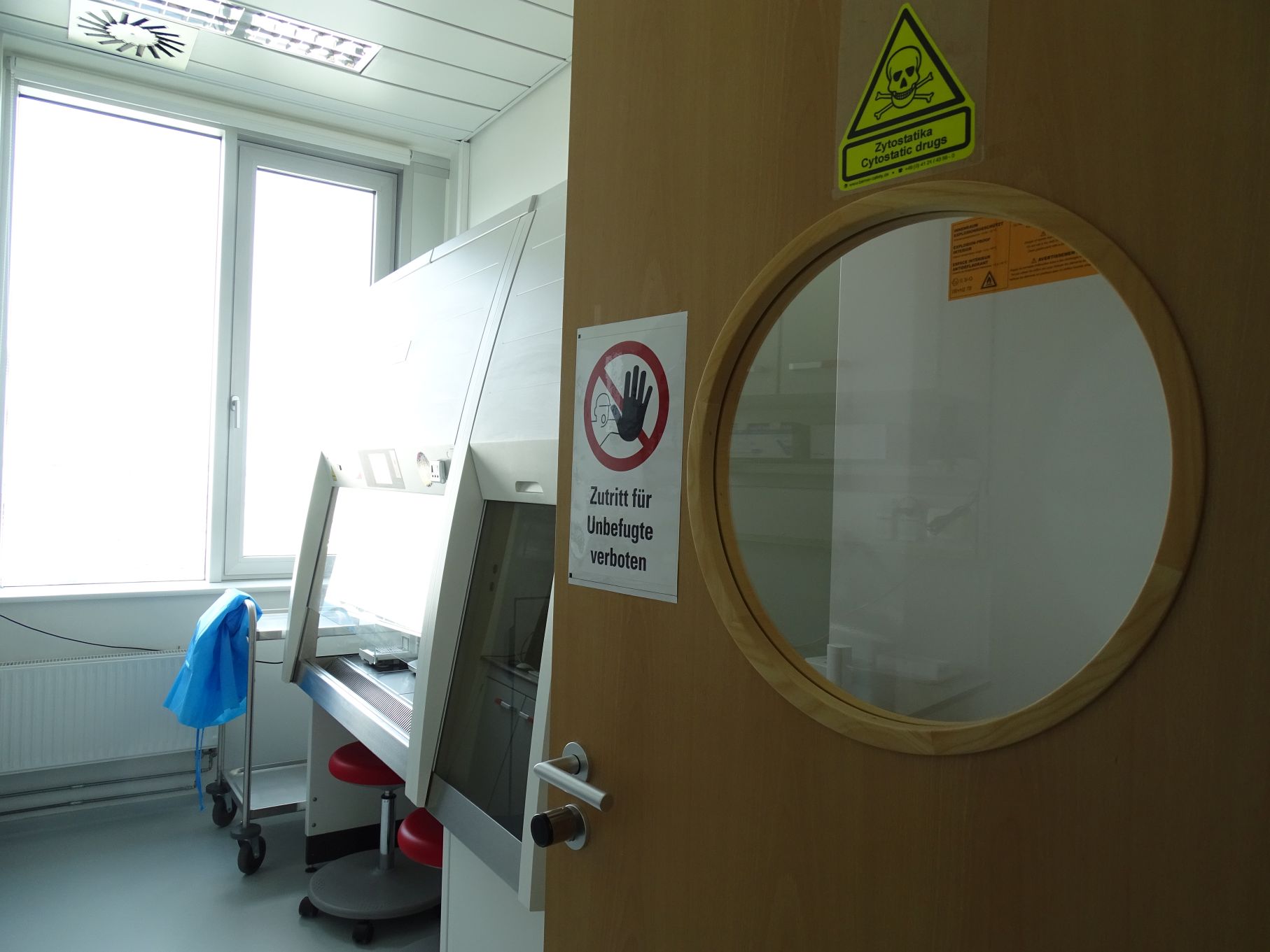 Auf dem Bild ist eine offene Tür mit Bullauge zu sehen. Auf der Tür sind zwei Warnschilder mit der Aufschrift „Zutritt für Unbefugte verboten“ bzw. „Zytostatika Cytostatic drugs“ aufgebracht. Durch die offene Tür sieht man eine Sicherheitswerkbank.