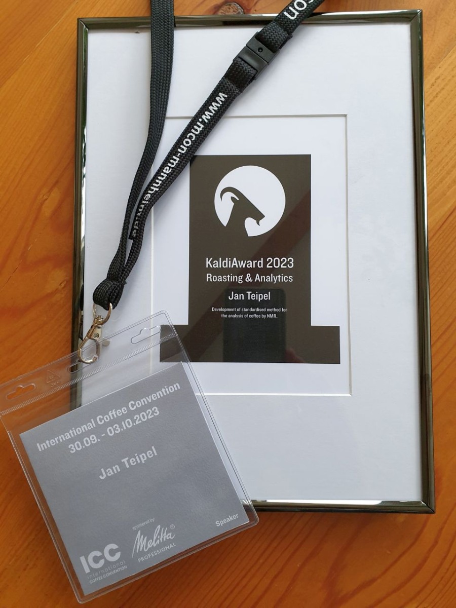 Die Abbildung zeigt den an Jan Teipel verliehenen, eingerahmten KaldiAward 2023 für besondere Leistungen auf dem Gebiet Röstung & Analytik und einen Vortragendenausweis für die International Coffee Convention vom 30.09. – 03.10.2023