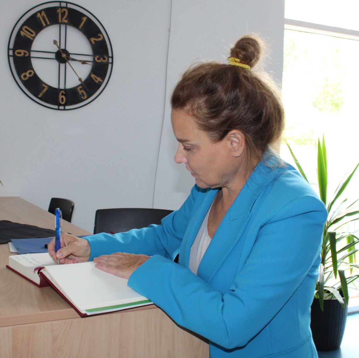 Eine Frau in leuchtend blauer Jacke steht an einem hohen Tisch und blickt in ein dickes Buch, in das sie schreibt.