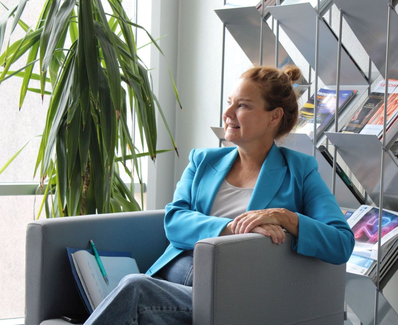 Eine Frau in leuchtend blauer Jacke sitzt lächelnd in einem grauen Sessel in einer Bibliothek. Sie hört und schaut aufmerksam zu.