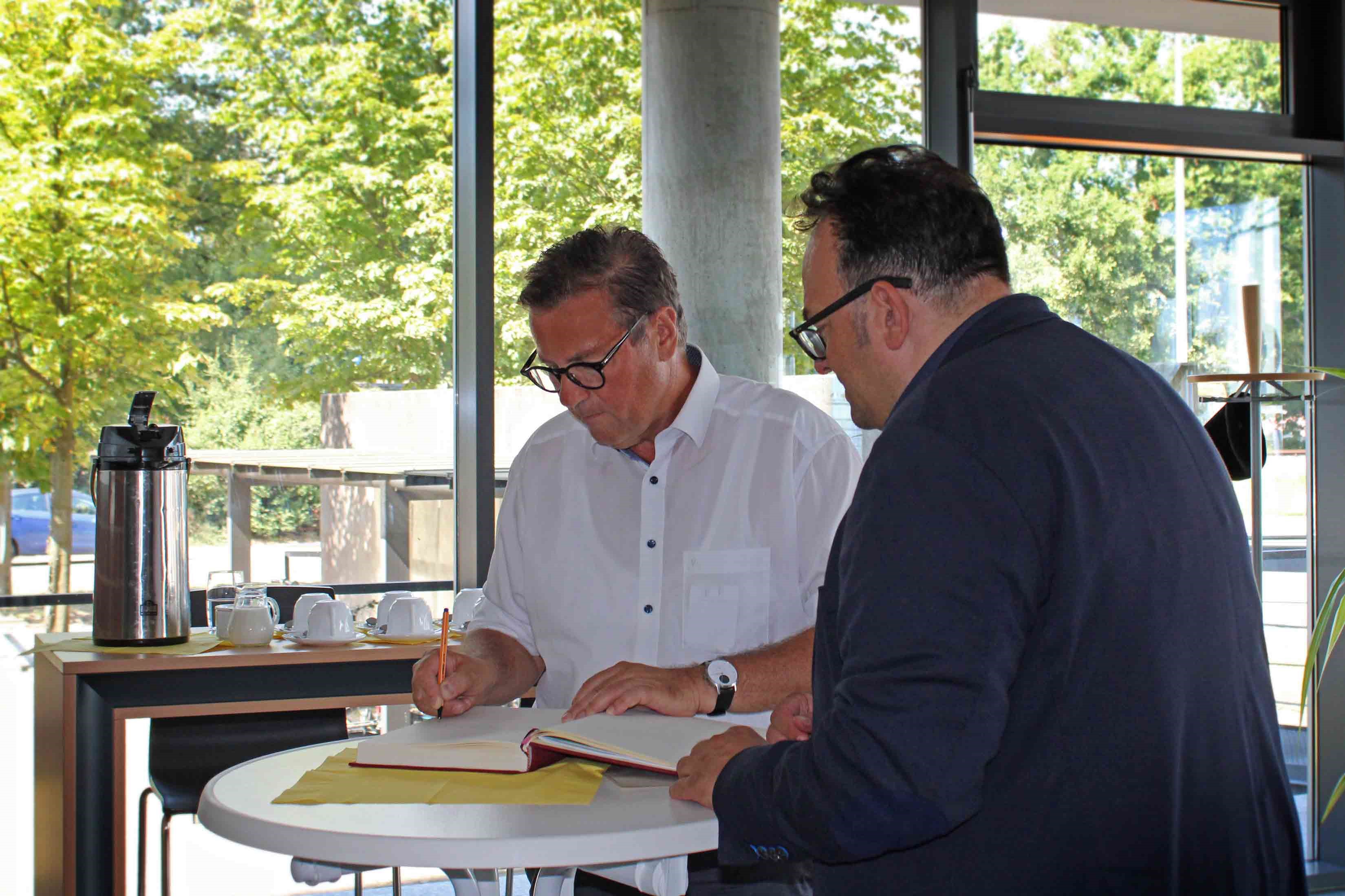 Auf dem Foto sieht man Herrn Minister Hauk und den Amtsleiter Dr. Walch gemeinsam an einem runden Tisch stehen. Der Minister trägt sich gerade in das Gästebuch ein.