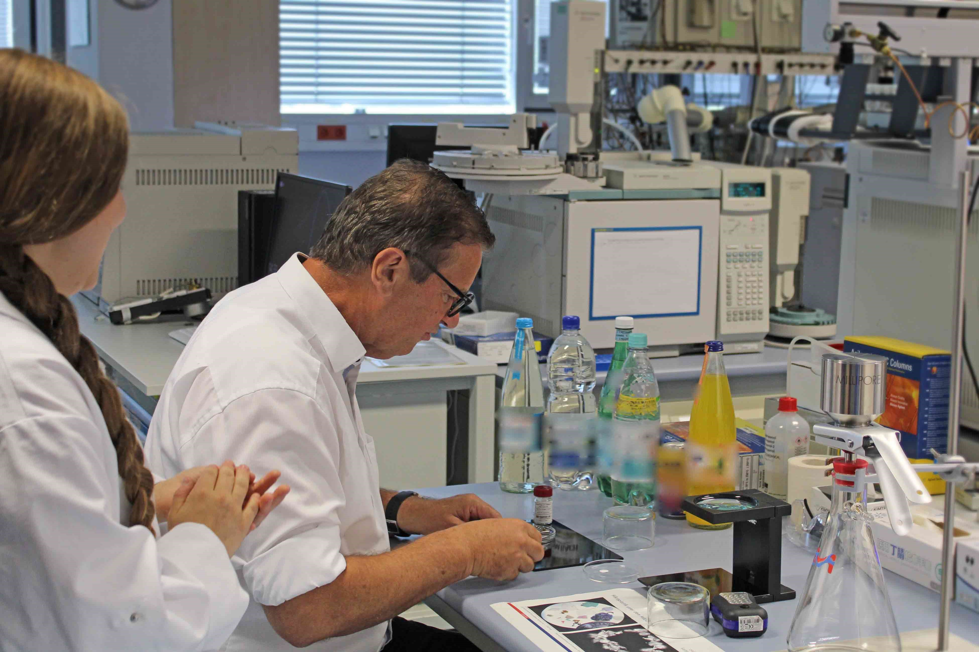 Auf dem Foto zu sehen ist eine Laborbank mit einigen Getränkeproben und einer großen Lupe. Der Minister sitzt davor und faltet (nicht zu sehen) einen kleinen Filter anhand den Erklärungen einer Sachverständigen, die neben ihm steht.