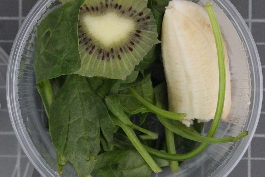 Das Bild zeigt in einer Kunststoffdose Spinatblätter, Bananen- und Kiwi-Stücke.