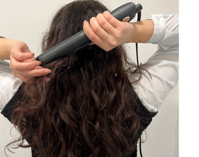 Das Bild zeigt eine Frau von hinten, die ein Glätteisen an ihre langen Haare hält.