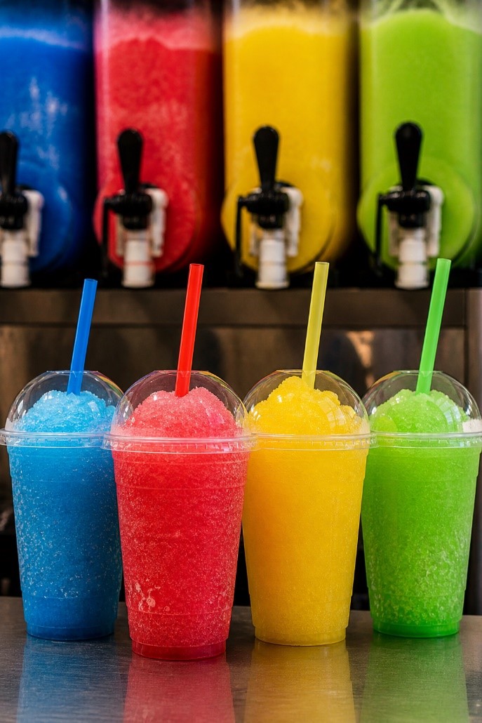 Das Bild zeigt vier bunte Slush-Eis-Getränke in Plastikbechern mit Strohhalmen. Dahinter stehen die zugehörigen Slush-Eis Maschinen.